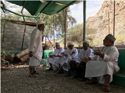 Five men in traditional white clothing sit and talk under a shaded canopy on a gravel area near a rocky hillside.