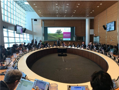 Large conference meeting in a modern room with attendees seated around a circular table, with a presentation screen at the front.
