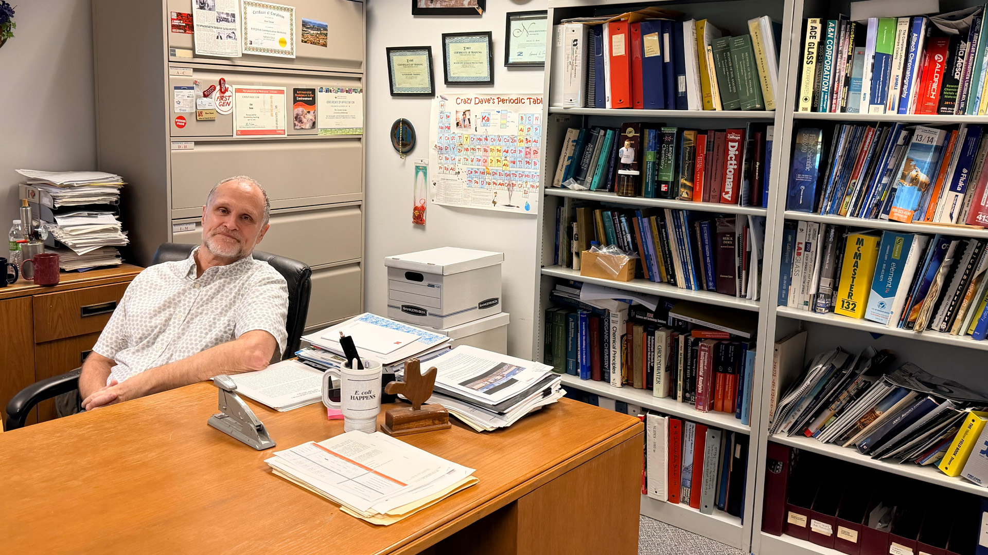 Man seated at a desk in a cluttered office, with tall bookcases of textbooks and binders and a periodic table poster on the wall.