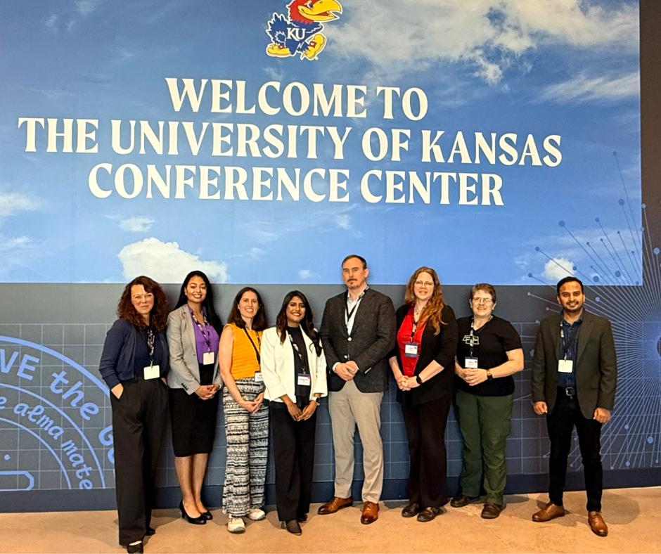 Group of eight conference attendees standing in front of a large sign reading “Welcome to the University of Kansas Conference Center,” with the KU Jayhawk logo above.