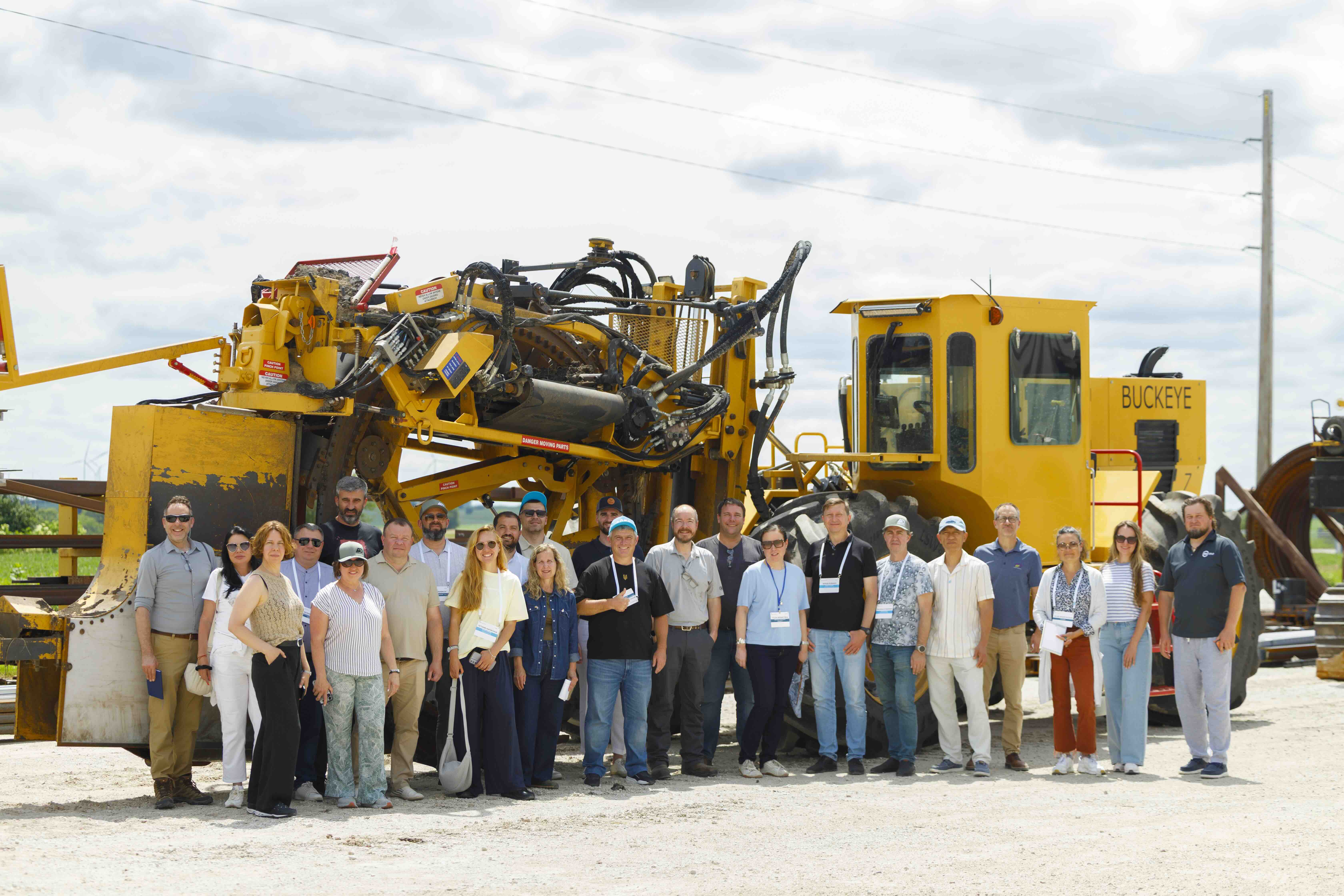 Group of people posing in front of a large yellow Buckeye road construction machine outdoors.