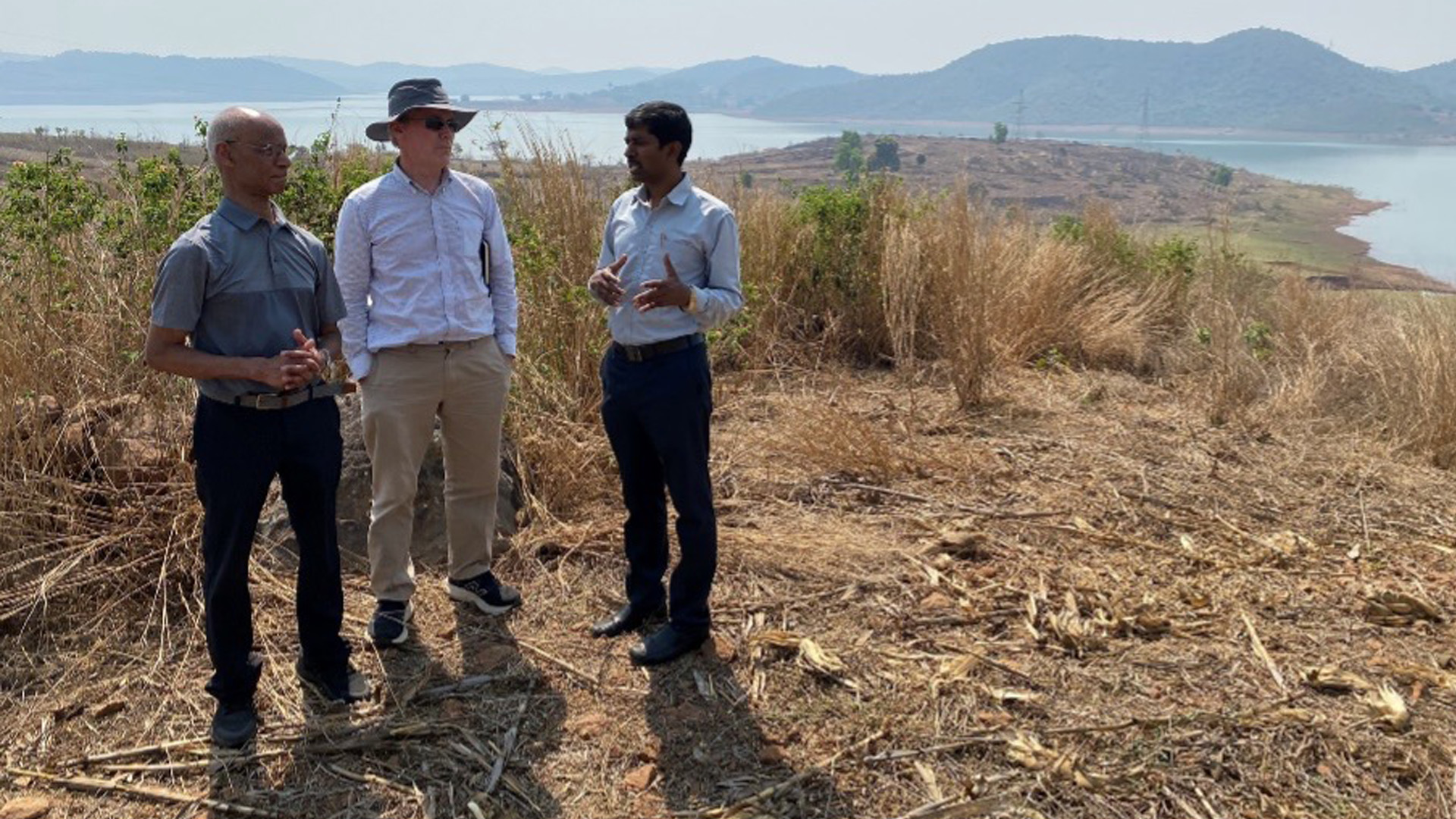 Three men stand on a dry hillside talking, with a lake and distant mountains in the background.