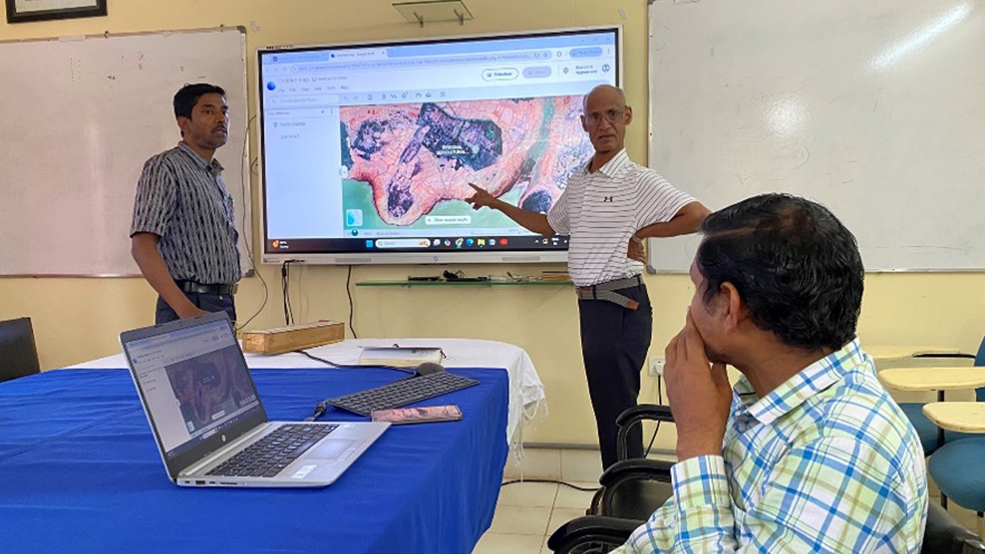 Two presenters in a classroom point to a satellite map displayed on a large screen while an attendee watches from a table with an open laptop.