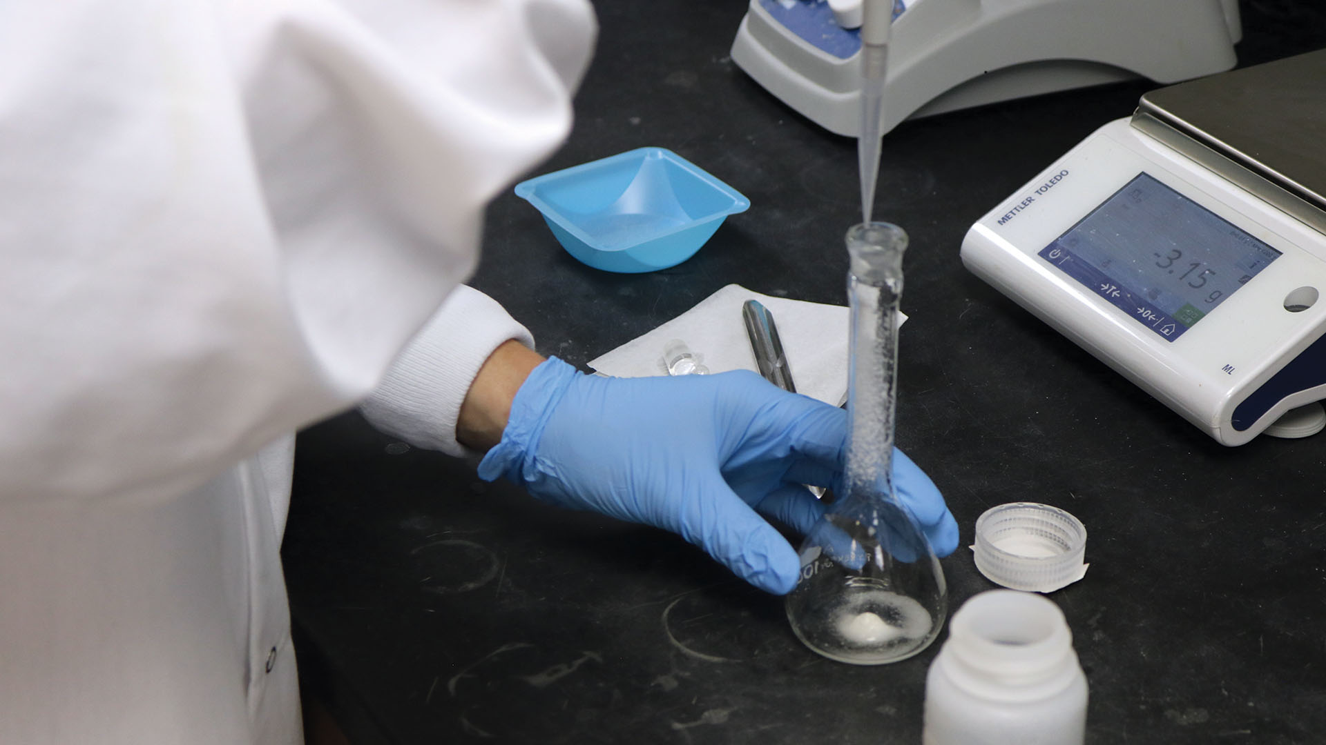 Gloved lab worker using a pipette to add liquid to a volumetric flask on a laboratory bench.