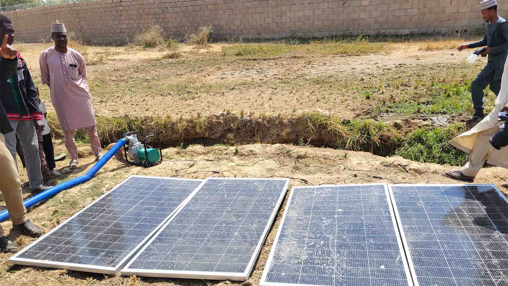 Solar panels powering a water pump with a hose, as people stand nearby in a rural field.